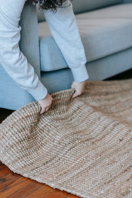 A person wearing a grey hoodie and blue pants is lifting the edge of a beige woven rug with a textured pattern, revealing a polished wooden floor beneath. The scene is set in a living room with a ligh