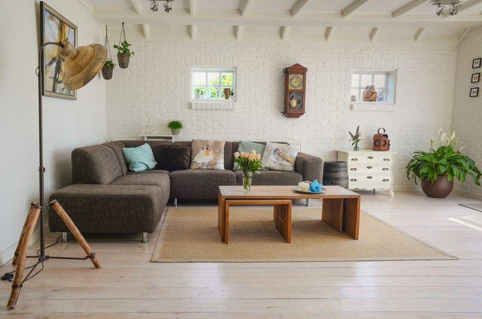 A person wearing a grey hoodie and blue pants is lifting the edge of a beige woven rug with a textured pattern, revealing a polished wooden floor beneath. The scene is set in a living room with a ligh
